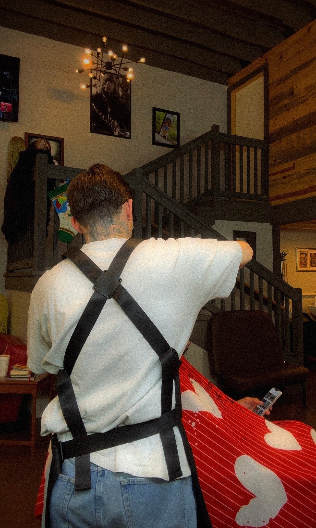 Person wearing white shirt and black harness standing indoors facing wooden staircase with wall decorations