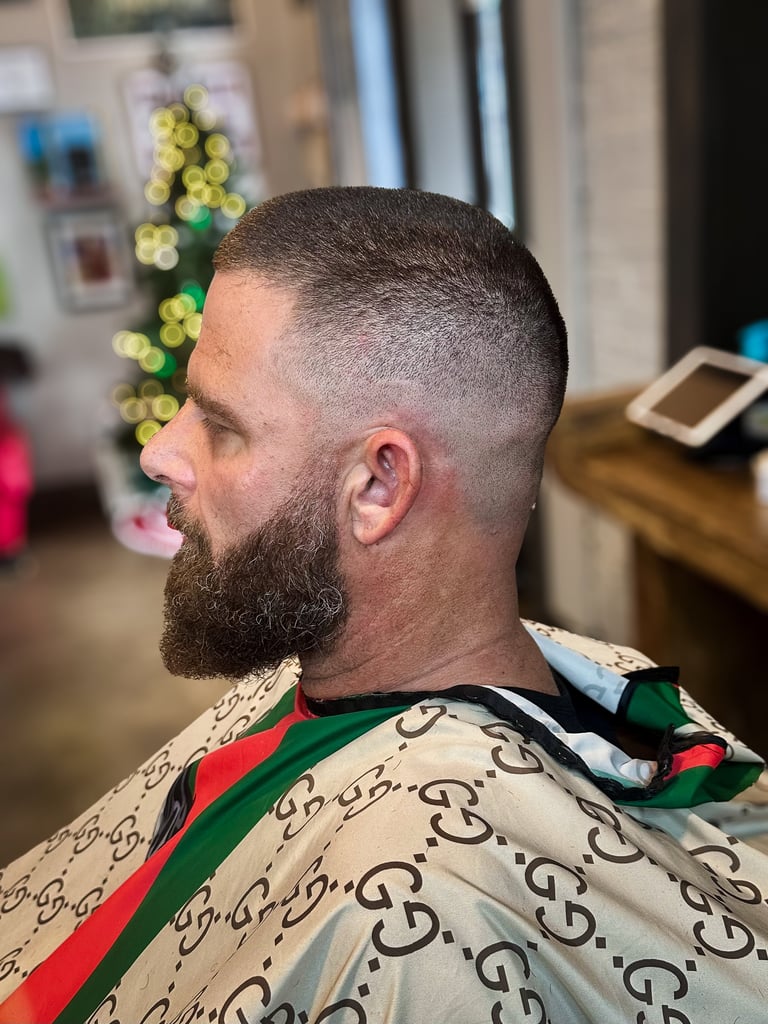 Man with short dark hair and full beard wearing patterned shirt, shown in profile view in a salon setting